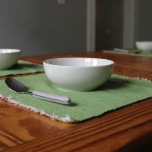 Green placemats with white bowls and grey handled spoons on a wooden table. Woven on a loom by Jacqueline's Textiles this green placemat is a handwoven fabric in NOVA. Green of the placemats is a light and fresh tea leaf or new leaf green.