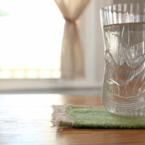 Light green coaster under clear glass coffee mug in front of natural handwoven bistro curtains. Mug rug in tea leaf green. Jacqueline's Textiles handwoven fabric in NOVA.