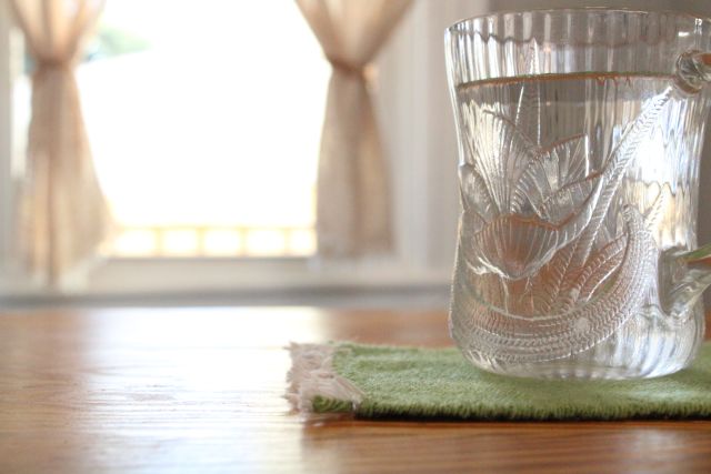Light green coaster under clear glass coffee mug in front of natural handwoven bistro curtains. Mug rug in tea leaf green. Jacqueline's Textiles handwoven fabric in NOVA.